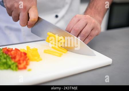 Crop man in weißer Chefjacke schneiden bunte Paprika mit Messer auf weißem Schneidebrett Stockfoto