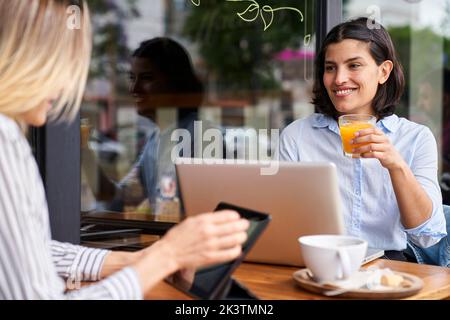 Mid-Shot-Foto einer lateinamerikanischen Unternehmerin, die Orangensaft trinkt, während sie mit ihrer Kollegin zusammenarbeitet Stockfoto