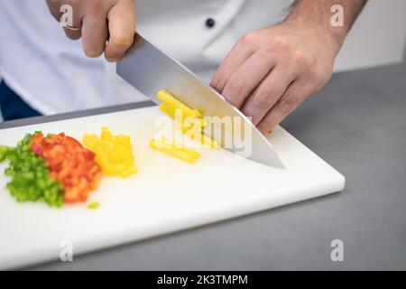 Crop man in weißer Chefjacke schneiden bunte Paprika mit Messer auf weißem Schneidebrett Stockfoto