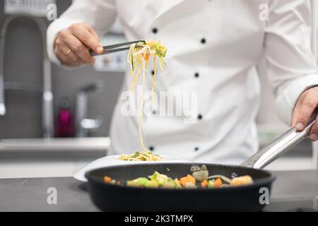 Crop anonymen Koch in weißen Uniform Kochen Pasta mit Gemüse in der Pfanne auf dem Herd auf dem Tisch in der hellen Küche Stockfoto