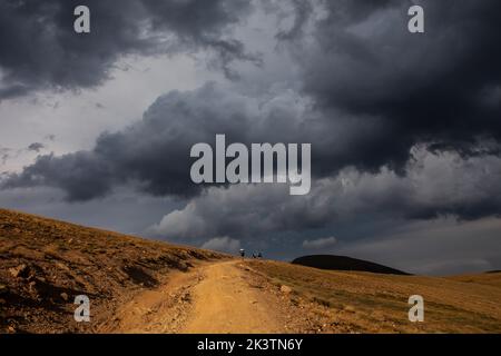 Entfernter anonymer Abenteurer, der auf einem sandigen Pfad durch hügeliges Wüstengelände unter einem düsteren, dramatischen Wolkenhimmel in den Pyrenäen geht Stockfoto
