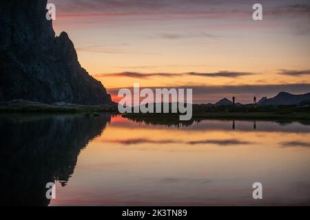 Friedliche Sommerlandschaft mit ruhigem See mit glatter Wasseroberfläche, die felsige Berge reflektiert und farbenfroh bewölktem Himmel bei Sonnenuntergang Stockfoto