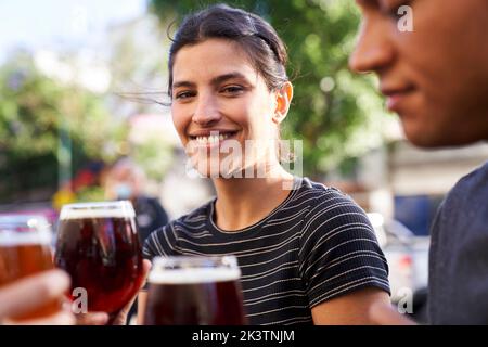 Porträt einer glücklichen jungen Lateinamerikanerin, die mit einem Lächeln auf die Kamera lächelt, während sie ein Glas Bier in der Hand hält Stockfoto
