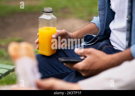 Mitten in der Hand eines Mannes, der eine Orangensaftflasche und eine unfokussiere Hand mit einem Sandwich hält Stockfoto