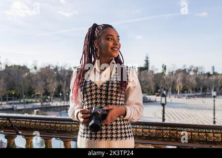 Lächelnde afroamerikanische Reisende mit moderner Fotokamera, die auf der Plaza de Espana steht und die Sehenswürdigkeiten während des Urlaubs in Sevilla genießt Stockfoto