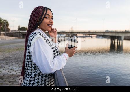 Seitenansicht einer lächelnden afroamerikanischen Frau mit Zöpfen und Kaffee, die in einer Papiertasse auf der Promenade steht und wegschaut, während sie die Abendstunden genießt Stockfoto