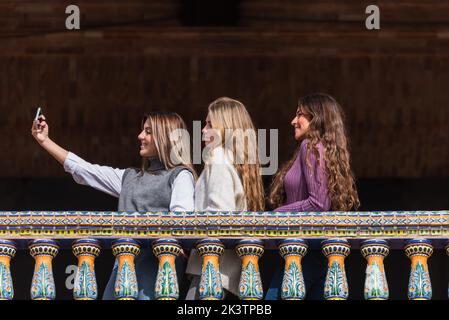 Flache Seitenansicht einer Gruppe von freundlichen Frauen, die am Wochenende in Sevilla auf dem Balkon auf der Plaza de Espana stehen und sich selbst auf dem Smartphone geschossen haben Stockfoto