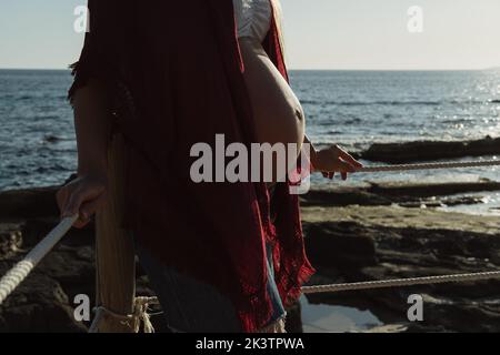 Beschnittenes, nicht erkennbares, schwanger Weibchen mit Hut, während sie in der Nähe des Zauns gegen Meer und blauen Himmel stand, während sie sich am Sommertag an der Küste ausruhte Stockfoto