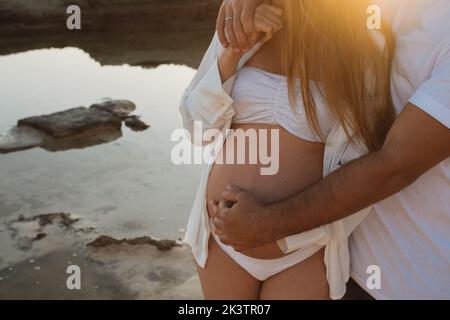 Ein unbekennbarer Mann, der eine Schwangere umarmt, während er morgens in der Natur an der Küste in der Nähe des ruhigen Meeres steht Stockfoto