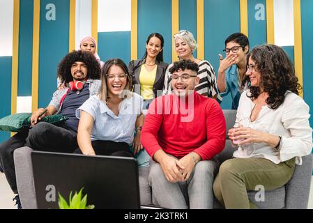Fröhliche, multiethnische Mitarbeiter in trendigen Kleidern sitzen auf einem bequemen Sofa und stehen, während sie sich Videos auf dem Laptop ansehen und im Modus gemeinsam lachen Stockfoto
