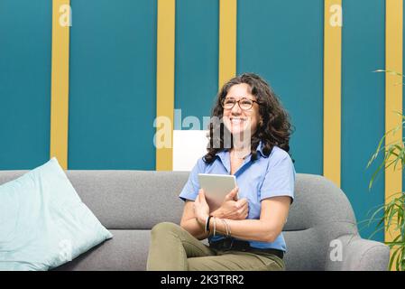Fröhliche reife hispanische Angestellte, die in der gemütlichen Lounge mit Tablet in einem modernen Arbeitsbereich in Costa Rica eine Pause einlegen und auf dem Sofa sitzen muss Stockfoto