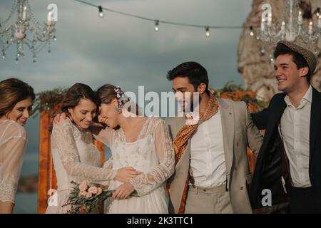 Junge lächelnde Jungs in Kostümen umarmt nahe attraktive fröhliche Damen in Kleidern mit Blumenstrauß in der Nähe von Dekorationen bei bewölktem Wetter Stockfoto