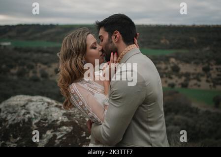 Rückansicht des jungen Mannes in Anzug umarmt mit Dame im Hochzeitskleid auf dem Gipfel des Berges und Blick auf die malerische Landschaft bei bewölktem Wetter Stockfoto