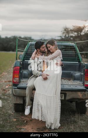 Junger Mann im Anzug sitzt in der Nähe attraktive Dame im Hochzeitskleid mit geschlossenen Augen auf männliche Schulter und sitzt auf Pickup-Truck Stockfoto