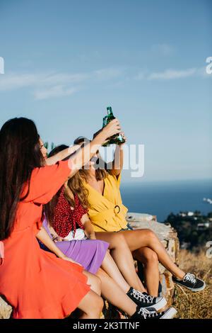 Eine Gruppe von unkenntlich fröhlichen Freundinnen in legerer Kleidung, die auf einer Steineinfassade sitzen und in klirrrenden Glasflaschen zusammenklirren, während sie zusammen Bier trinken Stockfoto