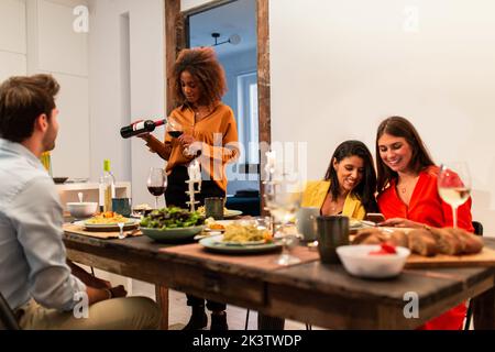 Fokussierte junge schwarze Frau, die Wein ins Glas gießt, während Freundinnen Handy benutzten und zu Hause zusammen zu Abend aßen Stockfoto
