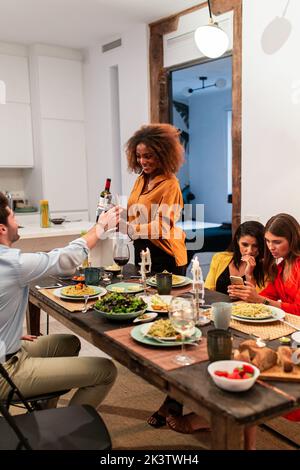 Fokussierte junge schwarze Frau, die Wein ins Glas gießt, während Freundinnen Handy benutzten und zu Hause zusammen zu Abend aßen Stockfoto