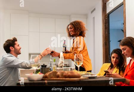 Fokussierte junge schwarze Frau, die Wein ins Glas gießt, während Freundinnen Handy benutzten und zu Hause zusammen zu Abend aßen Stockfoto