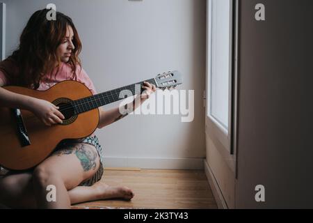 Junge rothaarige lachende Frau im Pyjama mit Gitarre, die auf das Fenster schaut und auf Treppen in der Dunkelheit sitzt Stockfoto