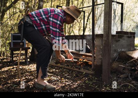 Ganzkörperexposition Argentinischer Mann mit Jagdhut und Leinenspadrillen, der im Wald Lagerfeuer aufhellt, während er im Wald Kessel kocht Stockfoto