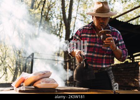 Älterer argentinischer Mann mit Schnurrbart, kariertem Hemd und schwarzer Hose, beigem Jagdhut und Wasserkocher in der Hand, der heißen Tee von t trinkt Stockfoto