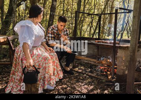 Argentinische Frau in traditioneller Kleidung mit Wasserkocher sitzt in der Nähe des Mannes trinken infundiertes Getränk gegen Fleisch, das über der Flamme auf dem Campingplatz gegrillt wird Stockfoto