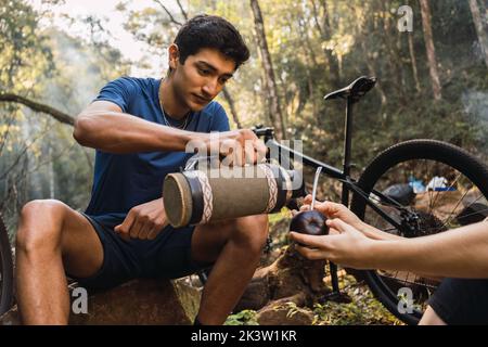 Seriöser ethnischer Mann, der auf Holzställen sitzt und während der Wanderung auf Fahrrädern im Wald in die Hände der Frau ein heißes Getränk aus Thermoskannen in anonyme Mate-Tasse gießt Stockfoto