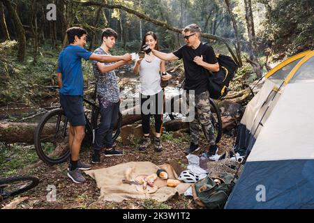 Ganzer Körper junger lächelnder multirassischer Freunde, die Tee aus Thermoskannen trinken, während sie in der Nähe des Zeltes im Wald stehen, während sie zusammen wandern Stockfoto