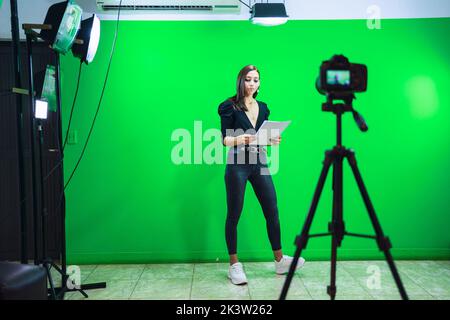 Ganzer Körper von weiblichen Moderatorin lesen Nachrichten auf Papier Dokument während der Aufzeichnung Programm für Social Media gegen Chromakey-Bildschirm Stockfoto