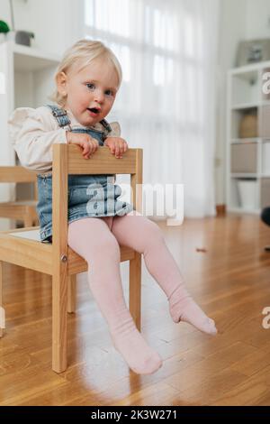 Niedliches kleines Kind in Denim-Overall-Kleid und Strumpfhose, das auf einem Holzstuhl sitzt und zu Hause wegschaut Stockfoto