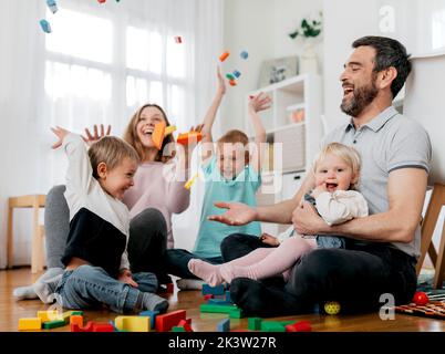 Fröhliche Eltern mit Kindern, die Modulblöcke in die Luft werfen, während sie im Lichthauszimmer spielen Stockfoto