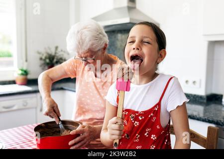 Sorgloses Kind mit Zunge aus und Spatel gegen Oma, die Teig für Kuchen in der Hausküche hastert Stockfoto