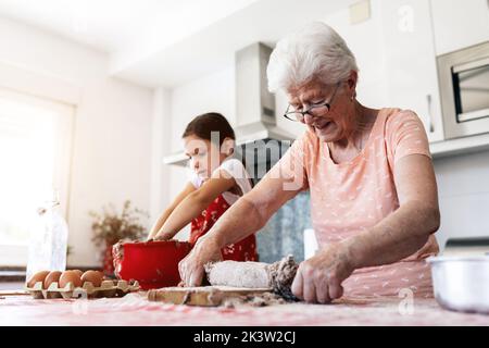 Fröhliche Oma in der Brille gegen die Enkelin mit Teig auf den Händen beim Kochen am Tisch in der Hausküche Stockfoto