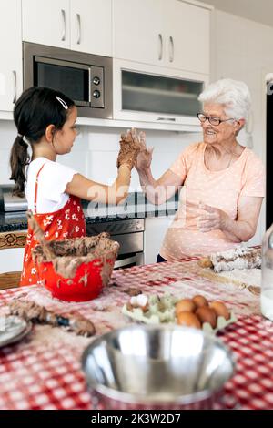 Fröhliche Oma mit Enkelin, die Spaß hat, während sie sich am Tisch mit Kuchenteig im Haus anschaut Stockfoto