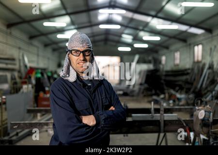Bärtiger Arbeiter mit schützendem Hardhut und blauem Overall, während er in der Garage steht und mit gekreuzten Armen auf die Kamera schaut Stockfoto
