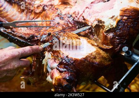 Der Koch schneidet das ganze gegrillte Schweinefleisch für Steaks mit einem Messer. Vom Schwein gegrillte traditionelle Kohle und Feuer. Das kleine Ferkel wird ganz auf einem offenen Feuer geröstet Stockfoto