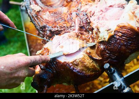 Der Koch schneidet das ganze gegrillte Schweinefleisch für Steaks mit einem Messer. Vom Schwein gegrillte traditionelle Kohle und Feuer. Das kleine Ferkel wird ganz auf einem offenen Feuer geröstet Stockfoto