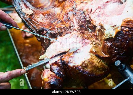 Der Koch schneidet das ganze gegrillte Schweinefleisch für Steaks mit einem Messer. Vom Schwein gegrillte traditionelle Kohle und Feuer. Das kleine Ferkel wird ganz auf einem offenen Feuer geröstet Stockfoto