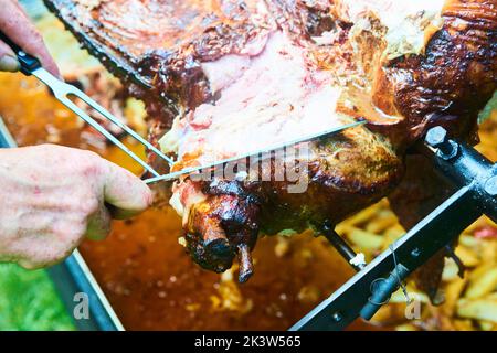 Der Koch schneidet das ganze gegrillte Schweinefleisch für Steaks mit einem Messer. Vom Schwein gegrillte traditionelle Kohle und Feuer. Das kleine Ferkel wird ganz auf einem offenen Feuer geröstet Stockfoto