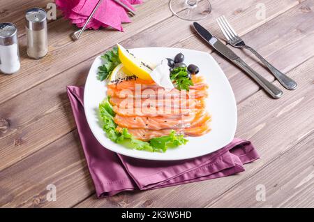 Frischer roter Fisch, der auf einem weißen Teller auf dunklem Holzgrund in Streifen mit Zitrone und Kräutern geschnitten wurde Stockfoto