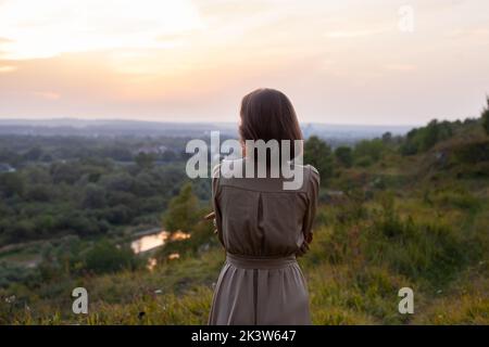 Schöne junge lächelnde Mädchen in einem langen braunen Kleid steht entlang des Rasens. Glückliche Frau geht bei Sonnenuntergang auf einem Hügel mit Blick auf den Fluss. Konzept des havi Stockfoto