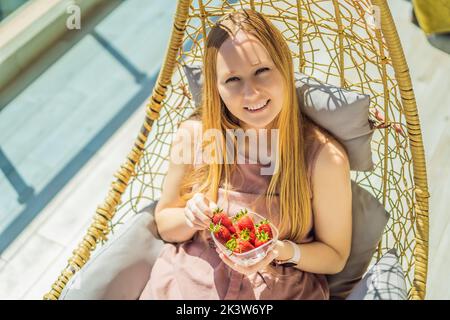 Portrait einer wunderschönen Frau in einem wunderschönen Kleid, die auf einer Terrasse sitzt und Erdbeere isst Stockfoto
