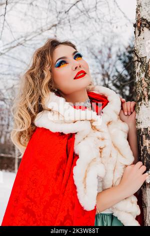 Ein Mädchen in roten Kleidern lehnt sich an einen Birkenwinterschnee Stockfoto