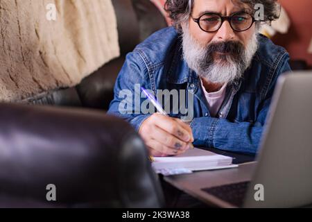 Mann auf einem Sofa mit einem Laptop zu Hause. Hipster notieren Sie sich beim Online-Lernen, und sehen Sie sich Webinar-Podcast auf dem Laptop-Hörkurs an, elearning Stockfoto