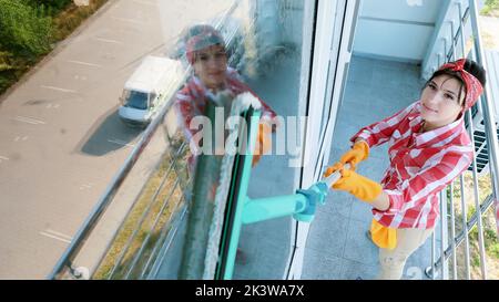 Im Fenster Glas reflektiert lächelnd kaukasische schöne Frau, in Handschuhen, Reinigung, Waschen Fenster durch spezielle Mopp, genießen ihre Arbeit. Reinigungsservice. Hochwertige Fotos Stockfoto