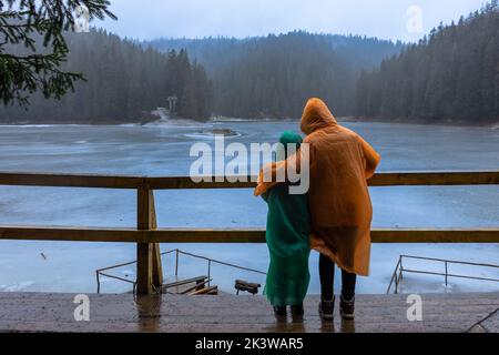 Zwei Kinder auf Regenmänteln genießen den mit Eis bedeckten See Synevyr, die Karpaten, Ukraine Stockfoto