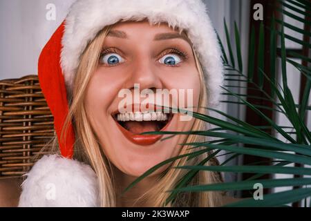 Lustige blonde Frau mit breitem Lächeln und Weihnachtsmütze, die mit überraschten Augen in die Kamera schaut. Mädchen feiert Weihnachten in den Tropen. Festliche Stimmung Stockfoto