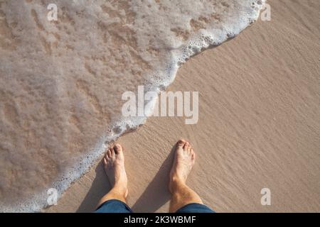 Barfuß Beine eines Mannes, der an einem sonnigen Tag in nassem Küstensand auf einem Strand steht, Draufsicht Stockfoto