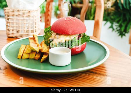 Veganer Burger, in einem roten Brötchen, Gericht mit Chips, auf einem grünen Teller, auf einem Holztisch, gesundes vegetarisches Essen. Stockfoto