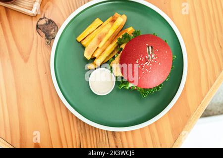 Veganer Burger, in einem roten Brötchen, Gericht mit Chips, auf einem grünen Teller, auf einem Holztisch, gesundes vegetarisches Essen. Stockfoto